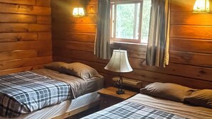 Ein Blockhauszimmer mit zwei Betten, Holzwänden, einem Fenster mit Vorhängen und einem Nachttisch mit Lampe.