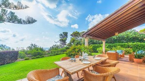 An outdoor patio with a wooden table and wicker chairs, surrounded by lush greenery and a clear sky.