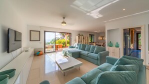 A modern living room with a large sectional sofa, a coffee table, and a TV mounted on the wall. There is a sliding glass door leading to an outdoor area with a patio table and chairs.