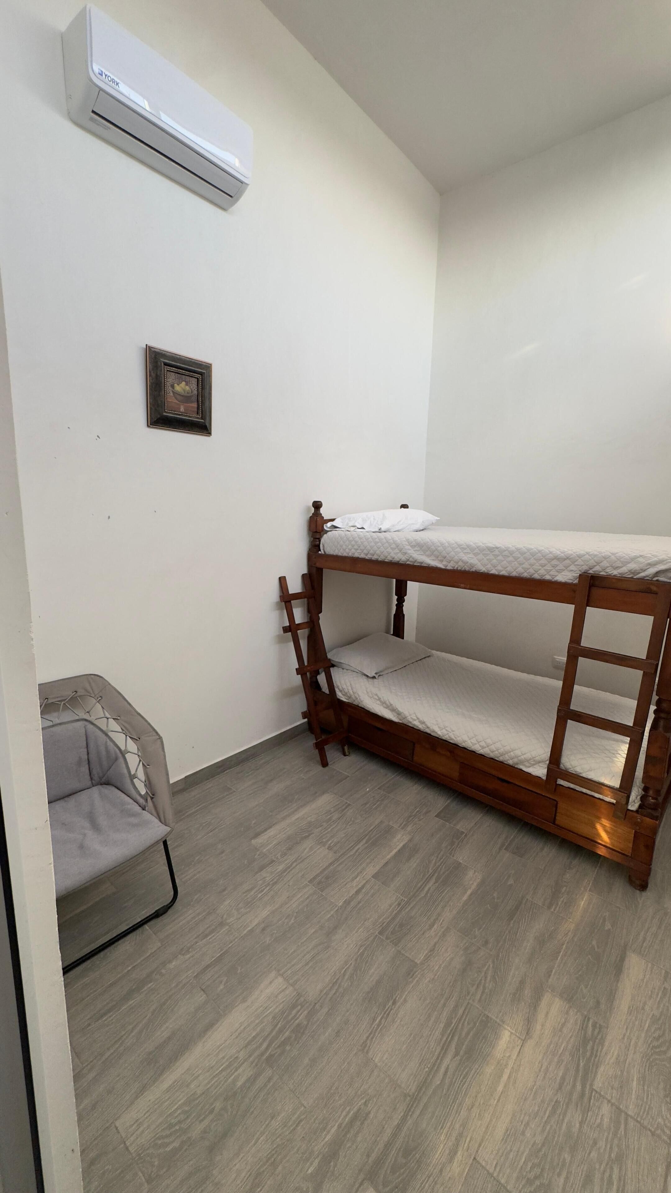 Een slaapkamer met een stapelbed met houten frame, een grijze stoel en een airconditioning aan de muur.