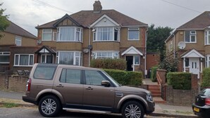 Un SUV marrón estacionado frente a una casa de dos pisos con un ventanal y una puerta principal.
