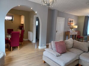 A living room with a sofa, a dining table with chairs, and a kitchen area visible through an archway. - Crawley Green House - Huku Kwetu (Luton)