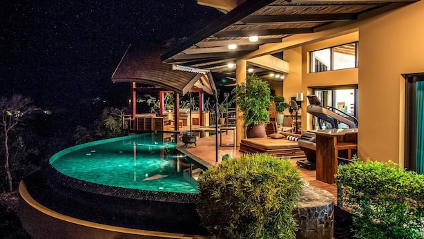 Zona de piscina exterior luxosa amb zona de seients coberta, una planta gran en test i vistes al cel nocturn.