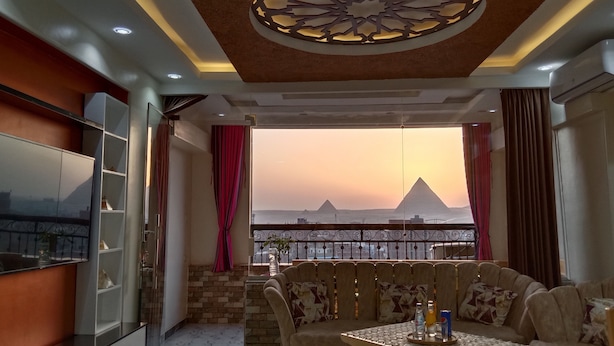 A modern living room with a view of the pyramids at sunset, a TV, and a balcony with a decorative ceiling. - Pyramids View apartment (Giza Governorate)
