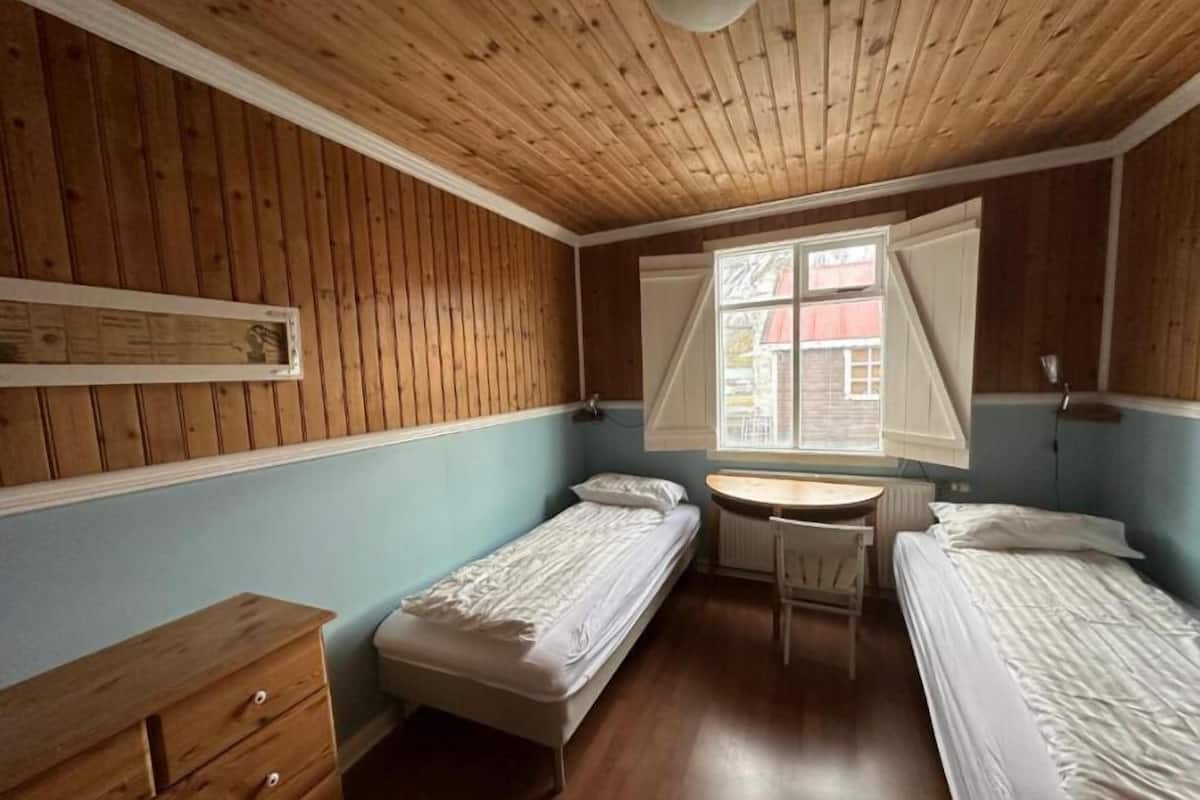 Une petite pièce agréable, avec des murs en bois et un plafond en pente. Elle comprend deux lits simples, un petit bureau en bois et une chaise.