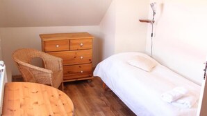 Een slaapkamer met een houten dressoir, een rieten stoel, een ronde houten tafel en een bed met witte beddengoed.
