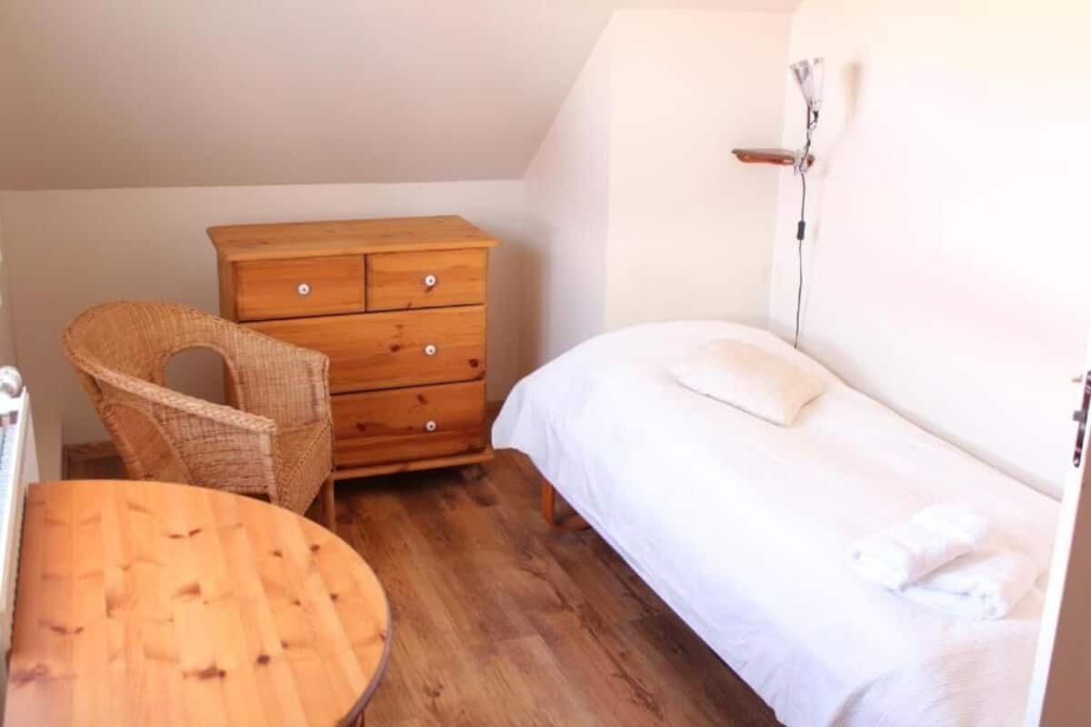 Une chambre à coucher avec une commode en bois, une chaise en osier, une table ronde en bois et un lit avec une literie blanche.