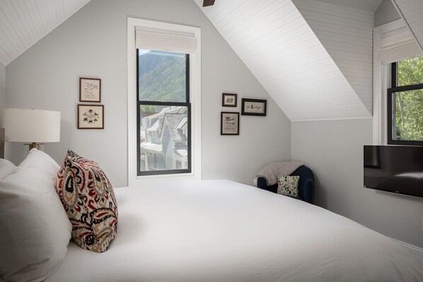 A bedroom with a large bed, a window with a view of a house, and a mounted TV.