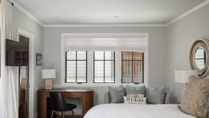 A bedroom with a bed, a desk, a chair, a window with blinds, and a mirror.