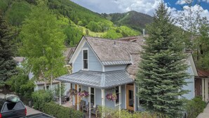 A two-story house with a porch, surrounded by lush greenery and a mountain backdrop.