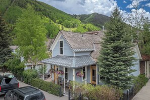 A two-story house with a porch, surrounded by lush greenery and a mountain backdrop. - The Residence on South Oak - Downtown convenience!, Perfect location by gondola and private hot tub! (Telluride)