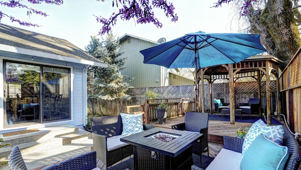 An outdoor patio with wicker furniture, a table with a fire pit, and a blue umbrella.