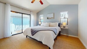 A bedroom with a bed, a ceiling fan, a window with a view, and a nightstand with a lamp.