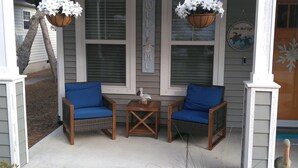 A porch with two wicker chairs with blue cushions, a small wooden table, and hanging flower baskets. - Sands About Right
 (Oak Island)