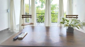 Ein Zimmer mit einem Holztisch, einer Fernbedienung und einer Vase mit weißen Blumen. Zwei Stühle und ein kleiner Tisch mit einer Pflanze stehen ebenfalls im Raum. Große Fenster mit weißen Vorhängen bieten Blick auf viel Grün.