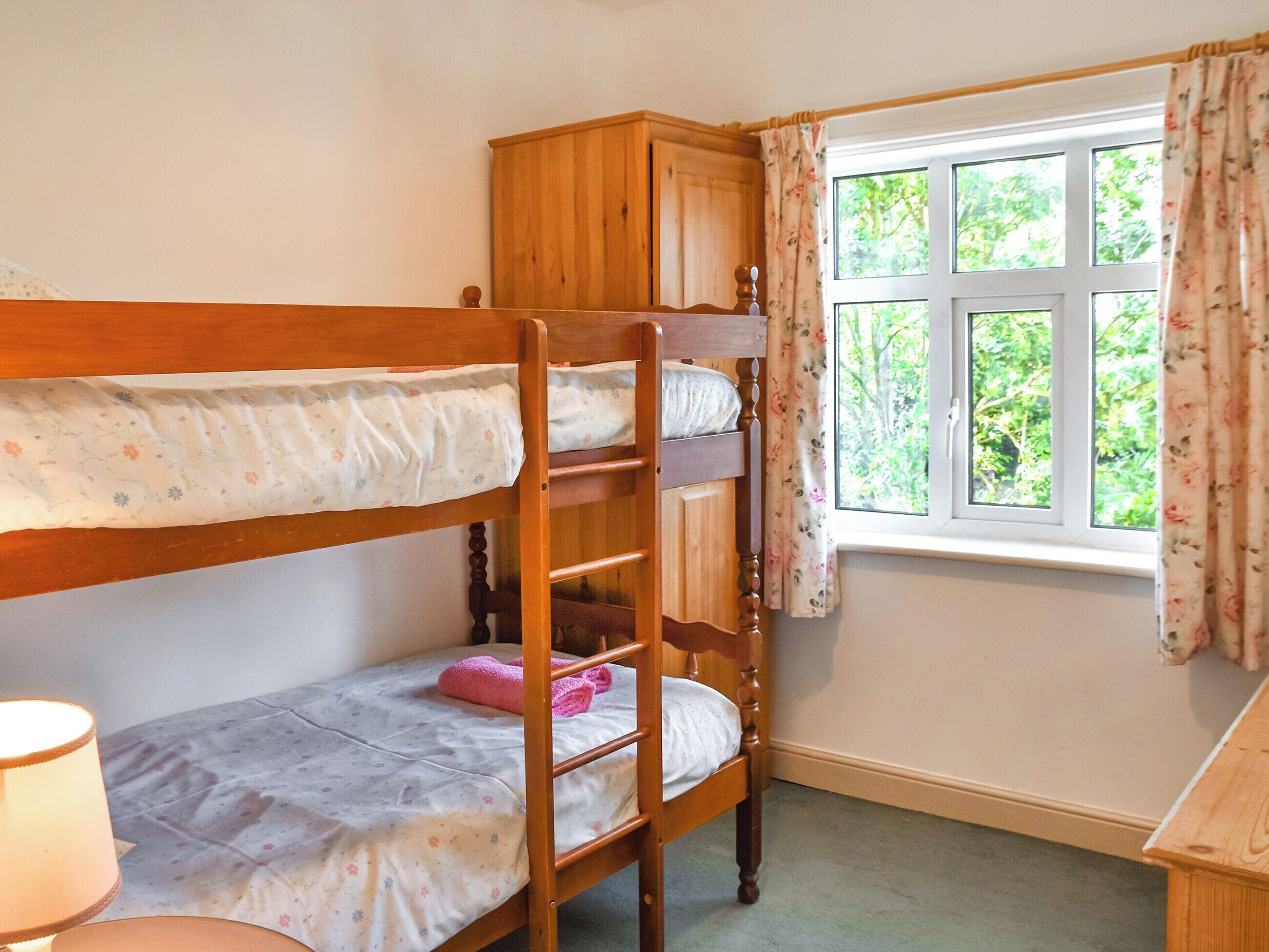 A bunk bed with a wooden headboard, a window with floral curtains, and a lamp on the left.