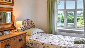 A bedroom with a bed, a wooden dresser, a lamp, a window with curtains, and a view of greenery outside. - 1 Brandiston Barn Cottage (Cawston near Reepham)
