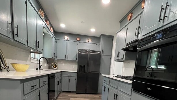 A kitchen with grey cabinets, a stainless steel refrigerator, a black oven, and a wooden floor.