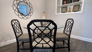 A dining area with a round table and chairs, a geometric wall mirror, and a shelf with framed pictures.