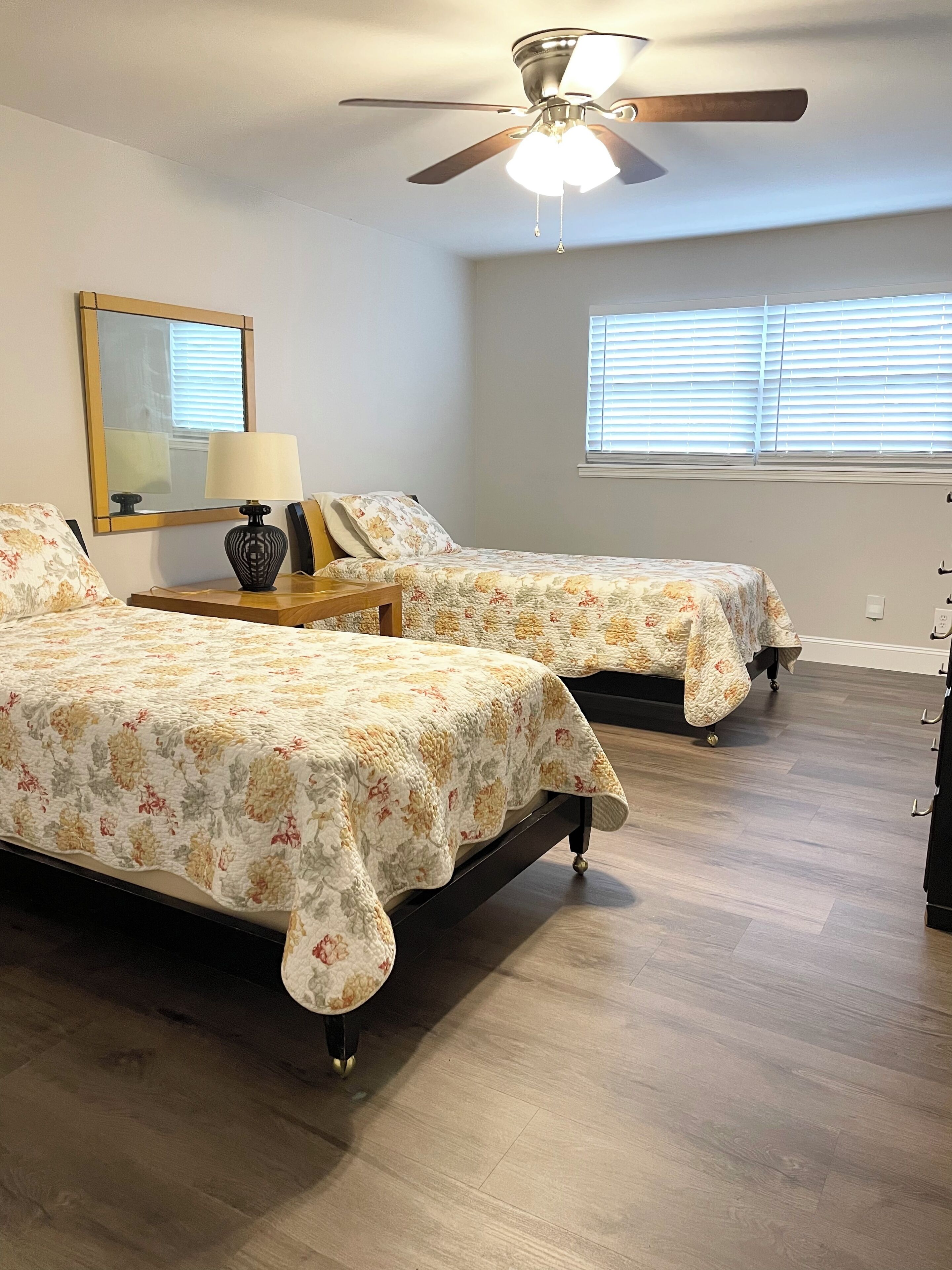 A bedroom with two beds, a ceiling fan, a nightstand with a lamp, a mirror, and a window with blinds.