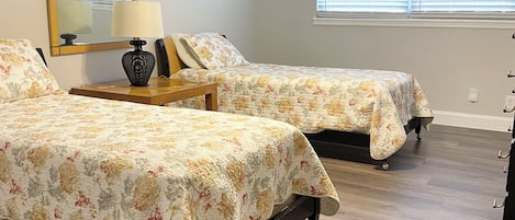 A bedroom with two beds, a ceiling fan, a nightstand with a lamp, a mirror, and a window with blinds.