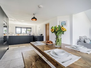 A modern kitchen with a central island, a dining area with a wooden table set for two, and a vase of sunflowers on the table. - The Old Coach Garage (Gerrans)