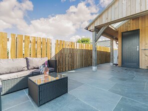 An outdoor patio with a sofa, a glass-top table, and a wooden fence. - The Old Coach Garage (Gerrans)