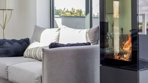 A modern living room with a sofa, cushions, a glass fireplace, and a large window. - The Old Coach Garage (Gerrans)