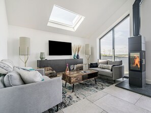 A modern living room with a fireplace, a flat-screen TV, and a large window. - The Old Coach Garage (Gerrans)
