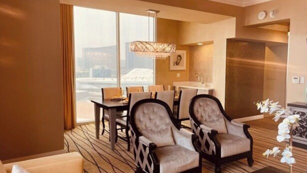 A hotel room with a dining area, two armchairs, a chandelier, and a view of a cityscape. - Four Bed Room Presidential Suite (Las Vegas)