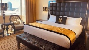 A hotel room with a large bed, a bench, a chair, a nightstand, and a view of the city. - Four Bed Room Presidential Suite (Las Vegas)
