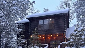 Une cabane en rondins dont le toit est recouvert de neige, avec une terrasse en bois et un escalier menant à l’entrée.