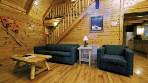 A wooden cabin interior with a staircase, two patterned sofas, a small table, and a kitchen area. - Cabins at Pine Haven (Beaver)