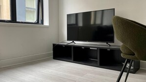Un salon moderne avec un téléviseur à écran plat posé sur un meuble noir, un fauteuil vert et une fenêtre équipée de stores.