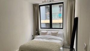 Une chambre à coucher avec un lit, une table de chevet, une lampe, une fenêtre avec des rideaux et une vue sur l’extérieur.