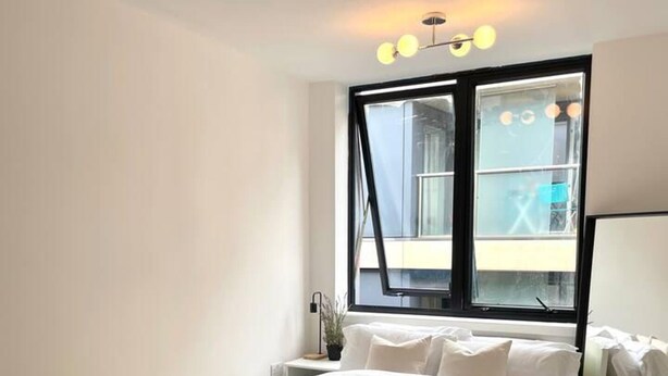 Une chambre moderne avec un grand lit, une table en verre, une fenêtre donnant sur l’extérieur et un luminaire au plafond.