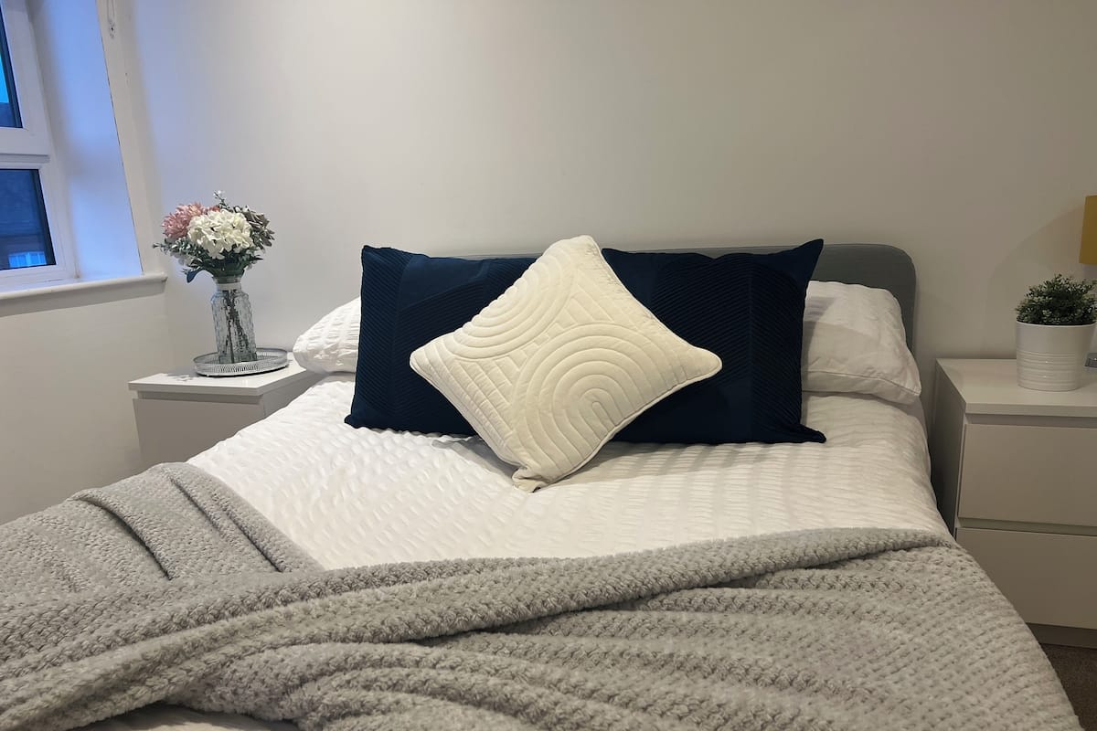 A neatly made bed with a white and navy blue pillow arrangement, a grey blanket, and a bedside table with a vase of flowers.