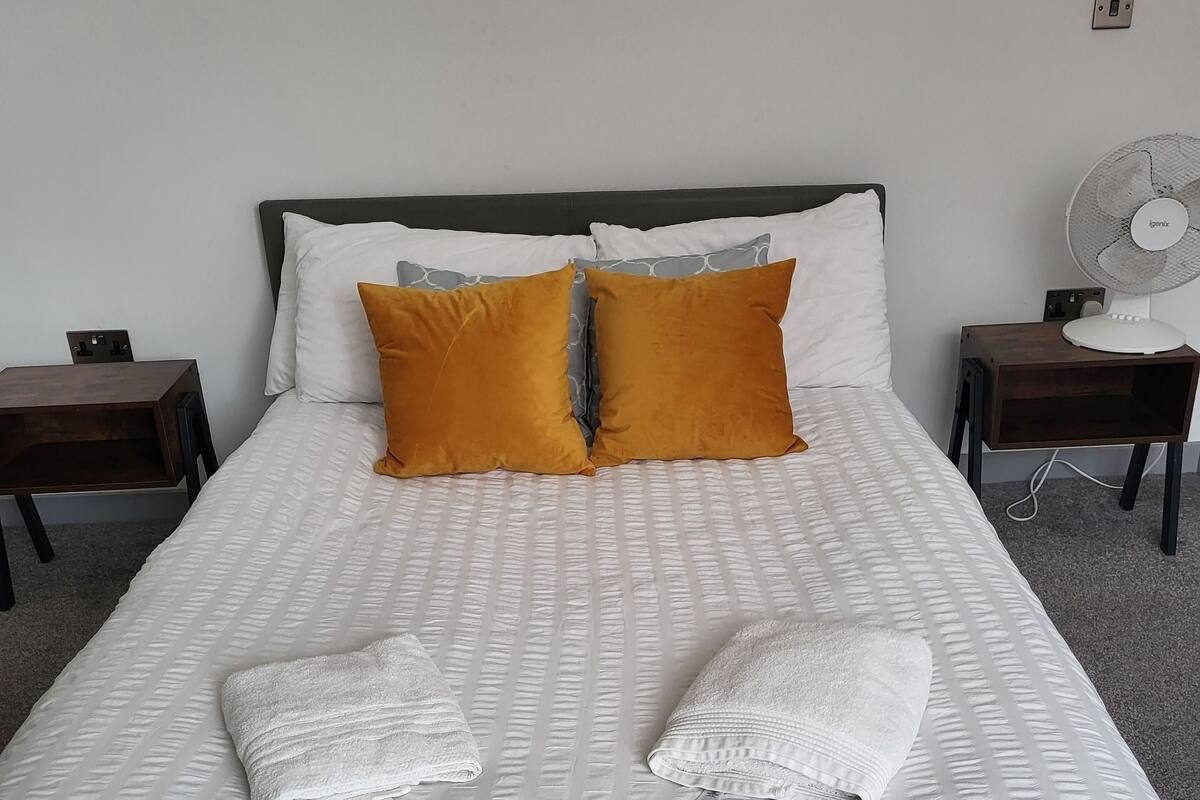 A neatly made bed with white bedding and two orange pillows, a bedside table with a lamp, and a wall-mounted fan.