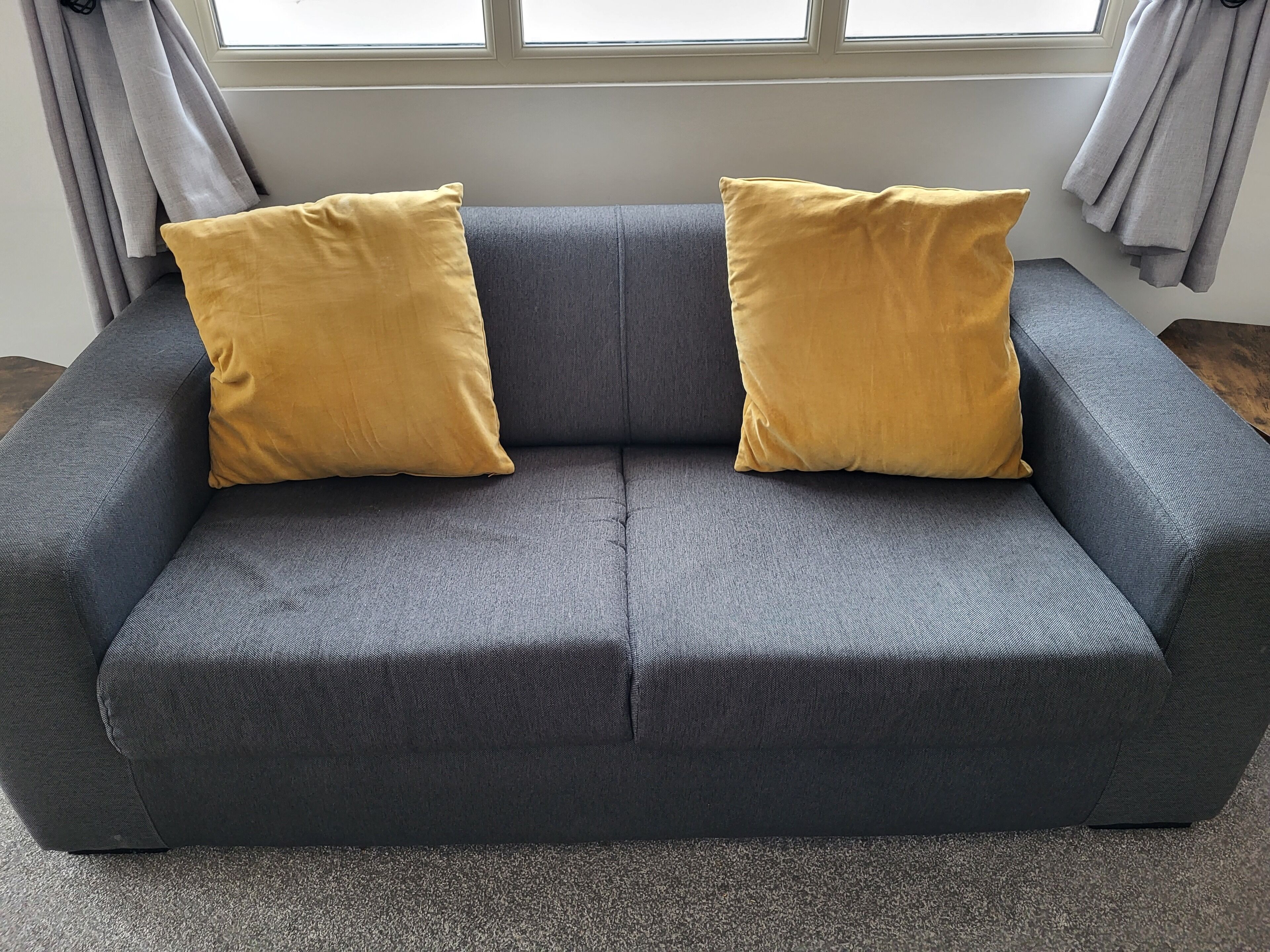 A grey sofa with two mustard yellow cushions, positioned in front of a window with grey curtains.