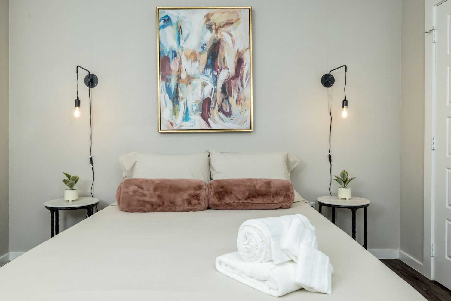 A bedroom with a large bed, two bedside tables with plants, a painting on the wall, and two wall-mounted lamps.