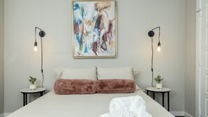 A bedroom with a large bed, two bedside tables with plants, a painting on the wall, and two wall-mounted lamps.