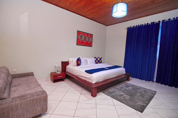 A bedroom with a wooden bed, a nightstand, a red bedside table, a grey rug, and a blue curtain. - The Haven (Ejisu)