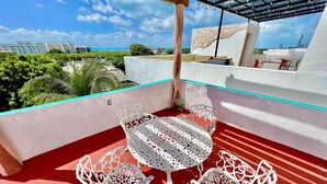 A rooftop terrace with white metal chairs and a table, a red floor, and a view of the ocean and buildings in the background. - Mid-Island Condo with Rooftop Balcony (Isla Mujeres)