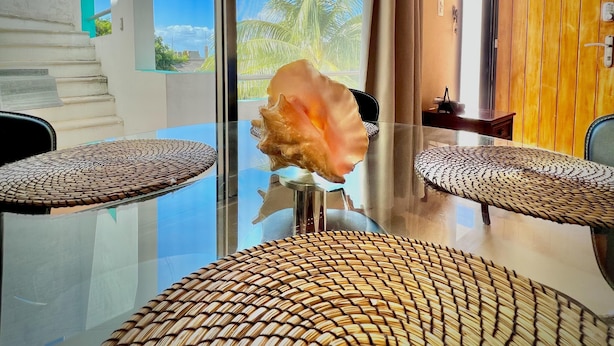 A modern room with a glass table, two chairs with woven seats, a large shell decoration, and a view of palm trees through the window. - Mid-Island Condo with Rooftop Balcony (Isla Mujeres)