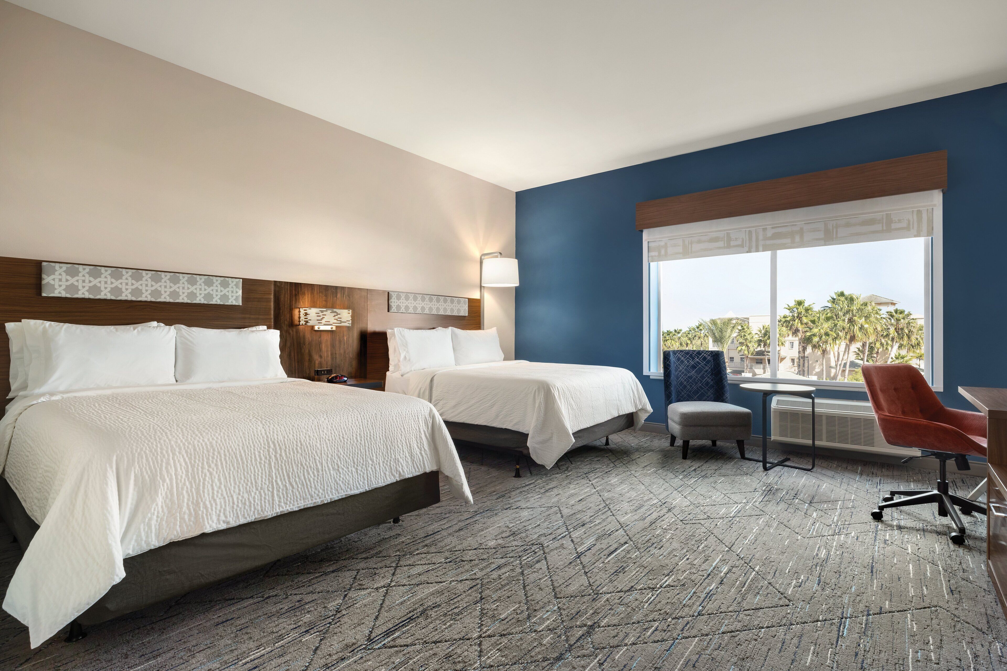 A hotel room with two beds, a desk, a chair, and a large window with a view of palm trees.