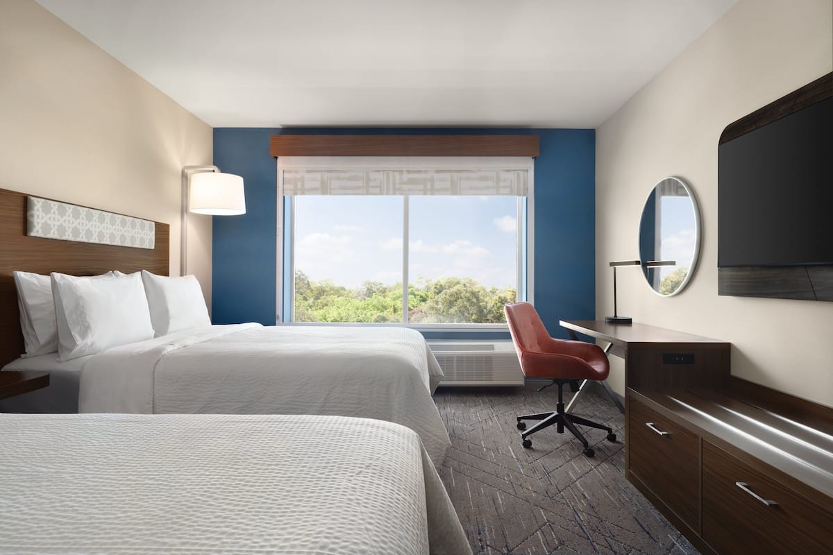Une chambre d’hôtel avec deux lits, un bureau, une chaise, une télévision et une grande fenêtre donnant sur un paysage verdoyant.