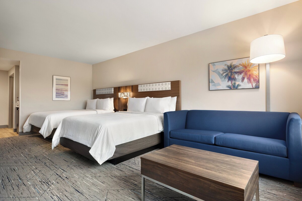 Ein Hotelzimmer mit zwei Betten, einem blauen Sofa, einem Holztisch und einem Palmenbild.