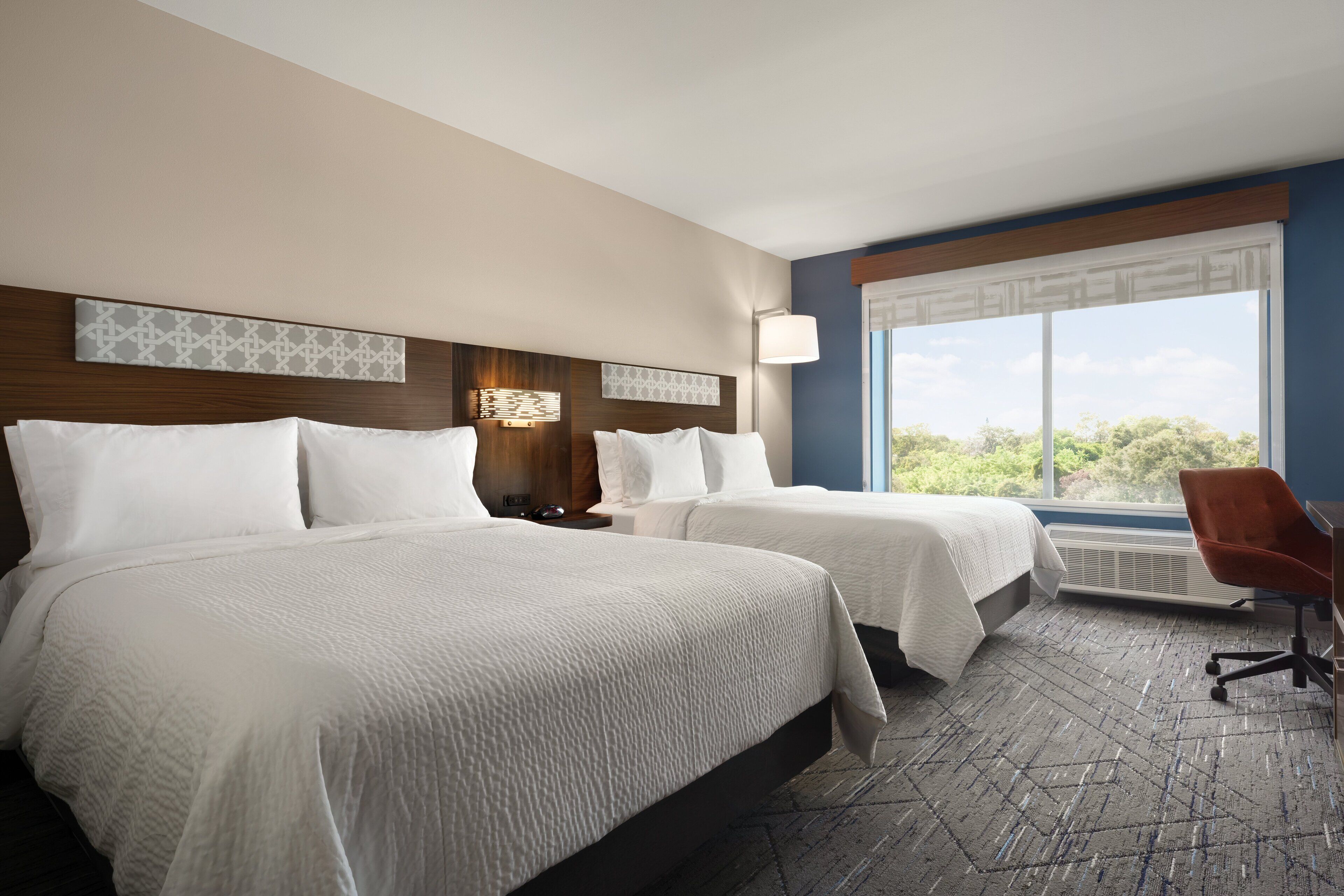 Habitación de hotel con dos camas, un escritorio, una silla y un amplio ventanal con vistas a zonas verdes.