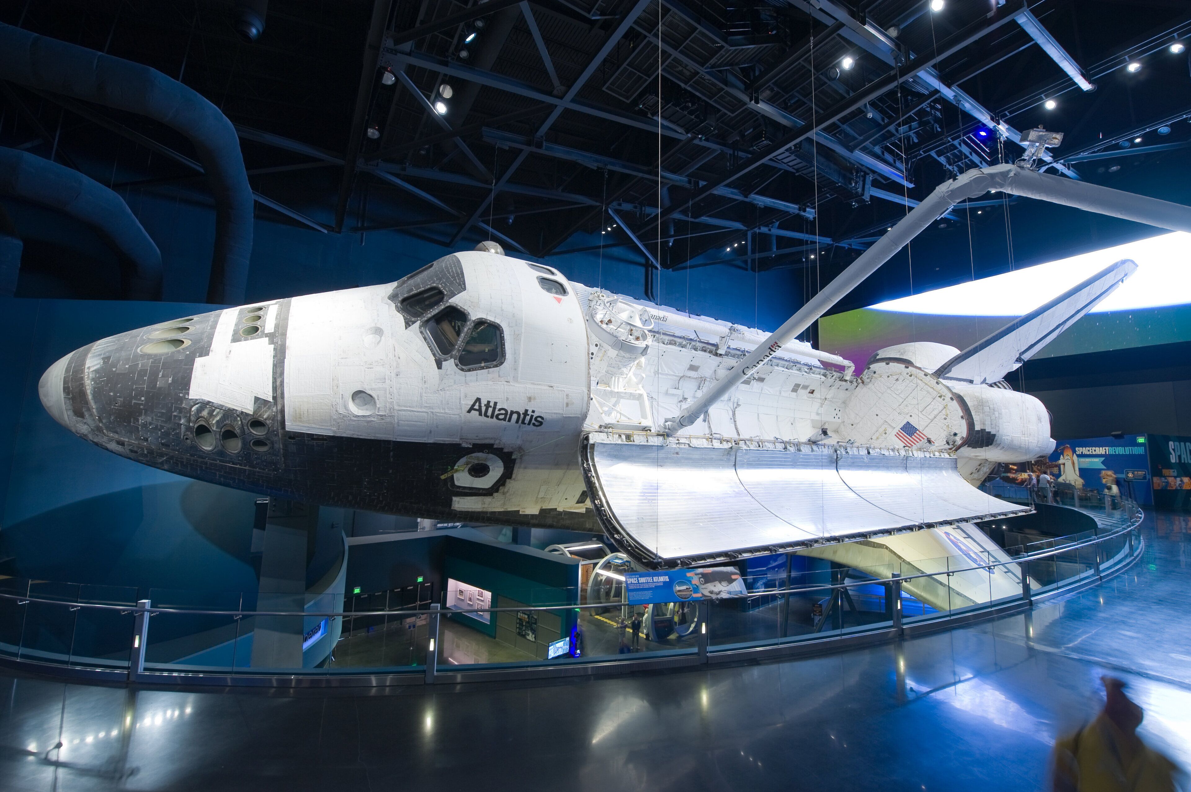 A space shuttle Atlantis displayed in a museum with a glass floor, allowing visitors to view the underside.
