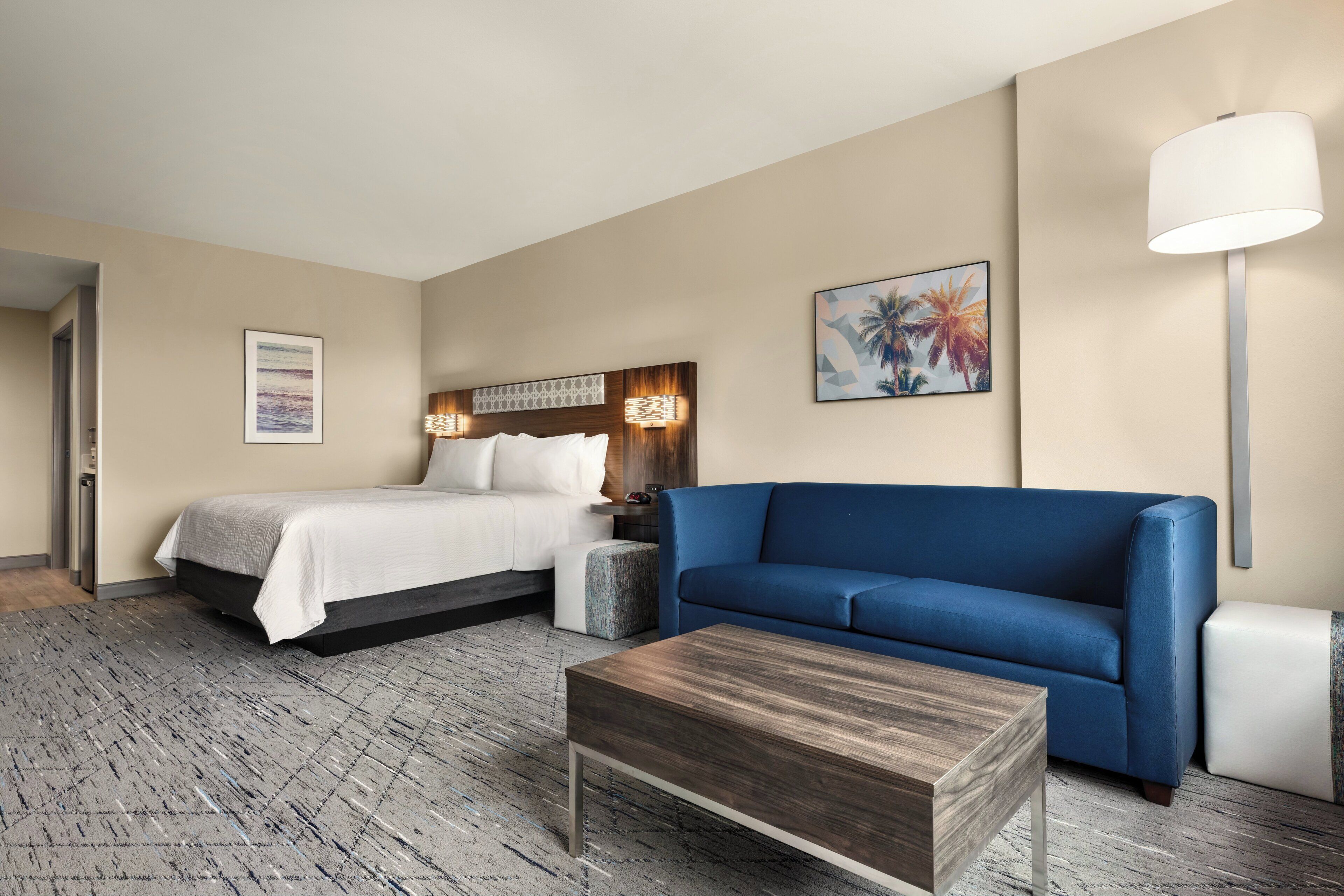 Una habitación de hotel con una cama, un sofá azul, una mesita de centro de madera y una mesita de noche con lámpara.